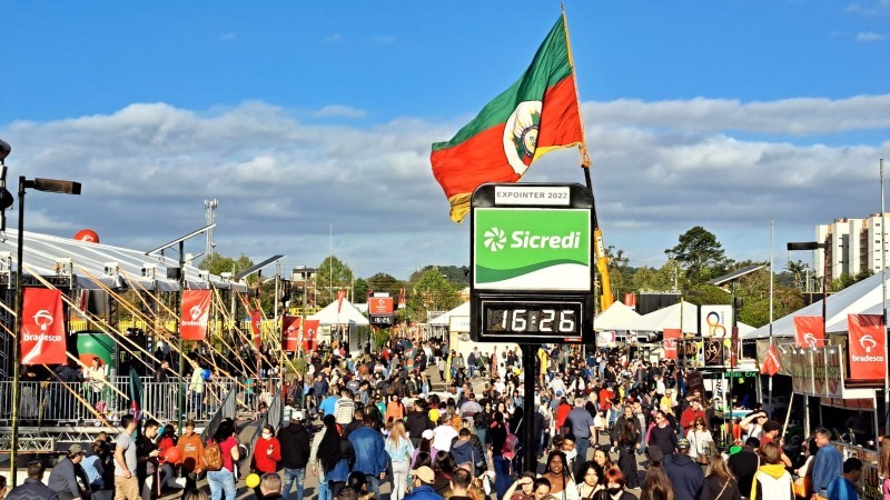 Foto de uma rua da Expointer. Céu azul com algumas nuvens, muitas pessoas visitando a feira. Nas laterais da rua os estandes e ao centro da imagem, no fim da rua, a bandeira do Rio Grande do Sul erguida por um guindaste. Também ao centro um relógio digital que marcava 16 horas e 26 minutos.