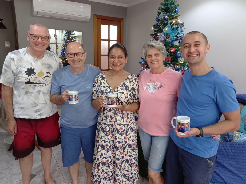 Foto do casal Arno e Clélia junto dos hospedes que receberam. Três homens e duas mulheres lado a lado, todos sorrindo. Cada um dos três hospedes receberam uma caneca de presente dos anfitriões e estão segurando-as.