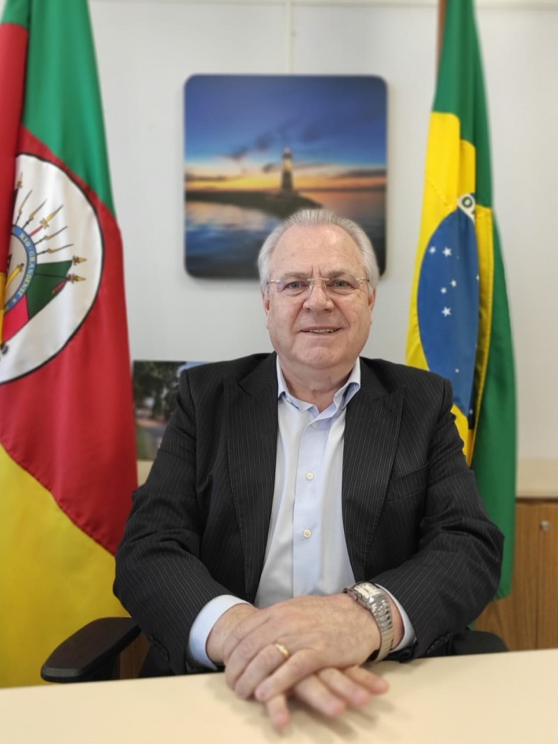 Secretário Vilson Covatti sentado ao centro, com a bandeira do Rio Grande do Sul a esquerda da foto, e a bandeira do Brasil a direita.