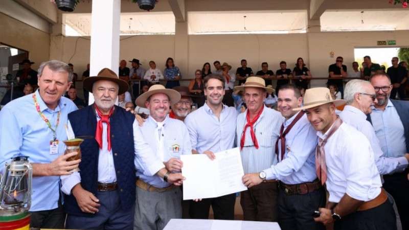Um grupo de homens sorridentes, muitos usando trajes t&iacute;picos ga&uacute;chos com len&ccedil;os e chap&eacute;us, posa segurando um documento aberto em um evento com p&uacute;blico ao fundo.